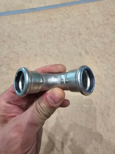 A hand holds a metal pipe fitting with two flared ends, each with a black rubber seal visible inside. The fitting is silver and cylindrical, with a slightly wider section in the middle. A faint green line is visible on the left side of the fitting. The background is a textured, light brown surface, possibly a table or floor, with some dark specks. A thin, light blue line runs horizontally across the top of the image. The scene is indoors, and there is no clear indication of the specific location in Darmstadt, Germany.