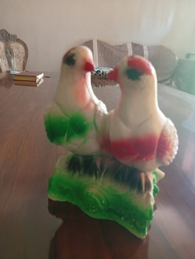 A figurine of two white birds with red and green accents is displayed on a wooden table. The birds are positioned side-by-side on a green base resembling foliage. In the background, a chair with an ornate back and a portion of a sofa are visible. The scene appears to be indoors. This image was taken in Akaltyn, Uzbekistan.