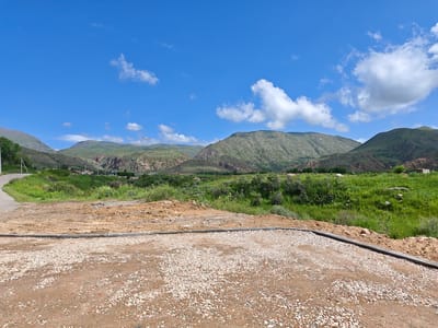 This is a landscape photograph taken on a clear day in Cholpon Ata, Kyrgyzstan. In the foreground, there is a rough, gravel-covered surface with some dirt. A concrete curb separates this area from a mound of brown dirt. Beyond the dirt mound, a grassy slope rises, dotted with patches of greenery and some scattered rocks. A paved road curves from the left side of the frame into the distance. In the background, rolling green hills and mountains form a picturesque backdrop against a bright blue sky with scattered white clouds. There are no people or animals visible in the image. The scene depicts a semi-developed area on the outskirts of a town, with elements of both natural landscape and human construction.