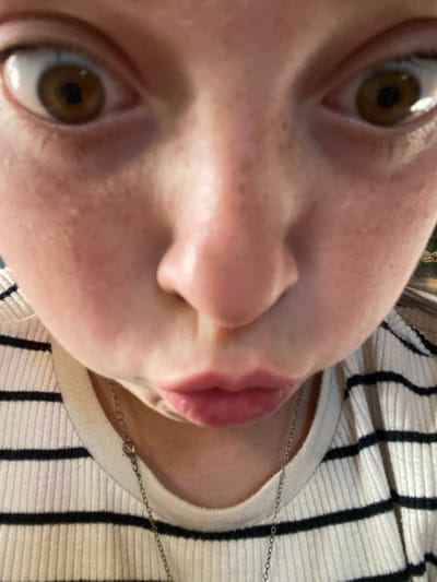 The image is an extreme close-up, low-angle, upside-down shot of a person's face, with the camera positioned very close to their nose and mouth. The subject has fair skin with visible freckles and brown eyes. Their mouth is slightly open, with the lower lip and tongue visible. A thin silver chain necklace rests on the person's chest, over a ribbed, cream-colored shirt with black stripes. The background shows a hint of trees and a bright sky, suggesting the photo was taken outdoors during the daytime. No text is visible in the image.