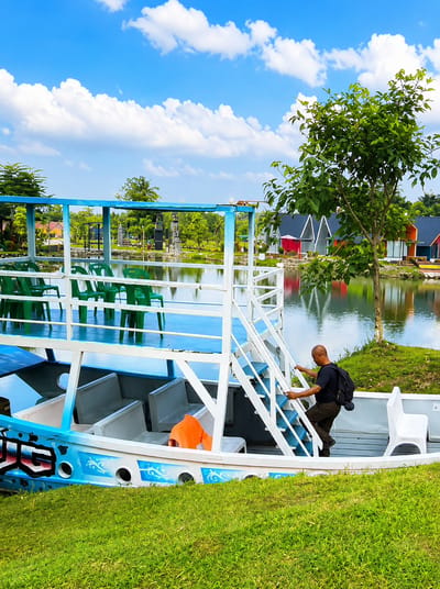 An exterior medium shot captures a man climbing a white staircase on the side of a blue and white boat. The man, wearing a black t-shirt and dark pants, has a black backpack on. He is positioned on the stairs, ascending towards the upper deck of the boat, holding onto the railing with his right hand. The boat features rows of white seats inside its hull, and an orange life vest is visible on one of the seats. The upper deck of the boat is painted blue and has several green chairs arranged in rows. In the background, a calm body of water reflects the sky. Across the water, several colorful buildings with distinctive A-frame roofs are visible, nestled amongst lush green trees. The sky is a vibrant blue, dotted with fluffy white clouds. The scene is set on a sunny day, with bright sunlight illuminating the environment. The location is Eampat Kwalamencirim, Indonesia, identified by the visible boat and the style of the buildings and landscape.
