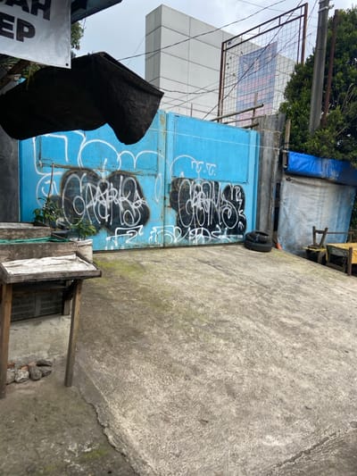 A large, blue, metal gate is painted with white graffiti. It is located in Lembang, Indonesia, at the coordinates -6.817, 107.613. To the left of the gate are two wooden stands and a green, metal fence. There are two discarded tires lying on the ground in front of the gate. There is also a large concrete structure that resembles a ramp leading up to the gate. To the right of the gate is a white, wooden structure partially obscured by a large, blue tarp. A tree stands in the background. The scene is partially obscured by a dark object in the foreground. The gate has multiple white graffiti marks that read "F.C." and other unreadable words.