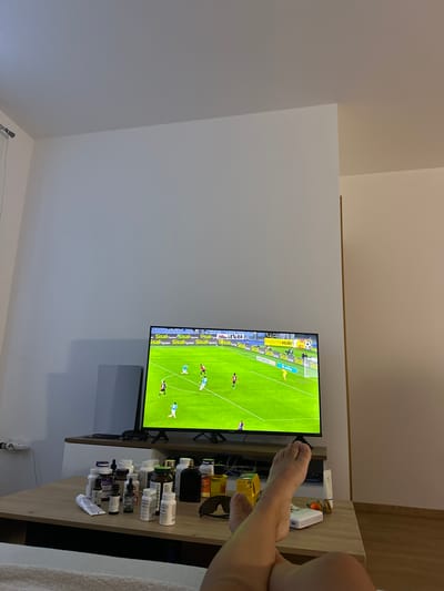 The image depicts a living room interior, likely in Donji Pobori, Montenegro, where a person is relaxing and watching a soccer match on a television. The television screen shows a live game with players on a green field and advertisements along the top border. In the foreground, a wooden table is covered with various bottles of supplements and medications, alongside a pair of sunglasses and other personal items. The person's legs and feet are visible, crossed and resting on the table, indicating a relaxed posture. The overall environment suggests a casual setting, possibly a home or an apartment.