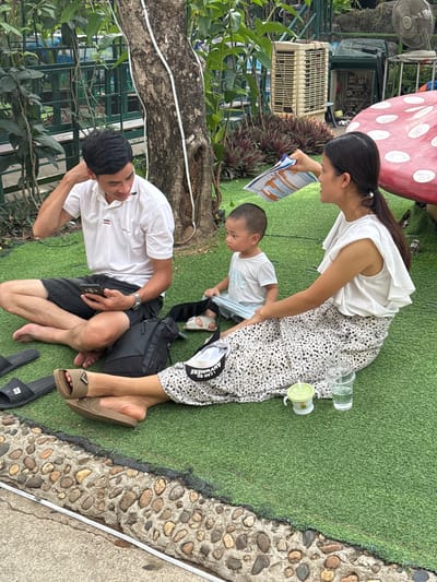 A family is having a picnic on artificial turf in the city of Hoa Lư Ward, Vietnam. The father, wearing a white polo shirt and black shorts, is sitting on the ground, looking at his phone. The mother, in a white ruffled blouse and a patterned skirt, is holding a brochure and interacting with their young son. The child, dressed in a striped t-shirt and blue shorts, is sitting between them. A large, red mushroom-shaped decoration is partially visible in the background, along with an air conditioning unit and some plants. Several pairs of sandals are scattered on the ground, and there are two drinks on the grass. The scene appears relaxed and casual, with the family enjoying their time together outdoors.