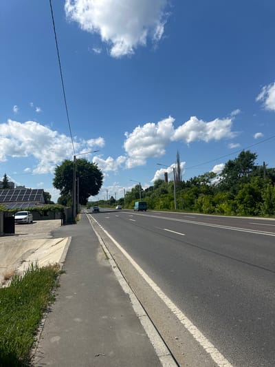 The image depicts a street scene in Strășeni, Moldova on a sunny day with a bright blue sky and scattered white clouds. The foreground shows a paved sidewalk with a grassy verge and a concrete drainage channel on the left. A single dark power line stretches diagonally across the frame from the top left.
The main focus of the scene is a two-lane asphalt road with white lane markings. Several vehicles are visible on the road: a light-colored car, a dark-colored car, a teal van, and two other cars further in the distance.
On the left side of the road, there is a house with solar panels on its roof, a fence, and a large, leafy tree. Beyond the house, more trees are visible. On the right side of the road, a dense line of trees and vegetation forms a natural backdrop. In the distance, industrial structures, including a chimney-like structure and tall poles, are present within the treeline. The overall environment suggests a semi-urban or suburban setting.