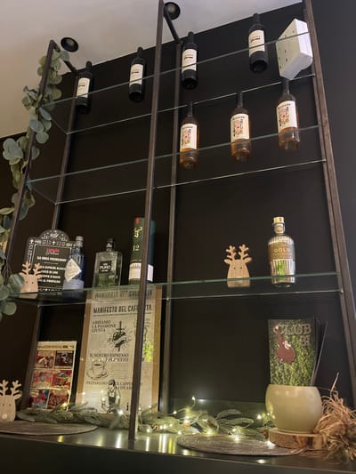 This image captures a section of a dark shelving unit or display case, likely in an indoor setting in Sondrio, Italy. The shelves are constructed from dark metal frames with transparent glass partitions, against a solid black background.
The display is primarily filled with various bottles. On the left, several wine bottles are arranged horizontally; some are dark-colored, while others are lighter with illustrated labels. On the right, various spirits bottles are visible, including one metallic gold bottle labeled "GOLD," a clear bottle marked "PURO," and a blue-tinted bottle with a tag mentioning "ESPOLON BLANCO." Another green bottle displays the number "12" and the partial word "fiddler."
Decorative elements suggest a festive or cozy atmosphere, possibly around a holiday season. These include several small, light-colored wooden reindeer cutouts placed on the shelves, artificial green leafy garlands (resembling eucalyptus) and pine branches intertwined with small string lights that emit a warm glow. A light-colored ceramic cup or candle holder with a rustic base of straw and wood is perched on the top right shelf. A white power strip is partly visible on the upper left shelf.
Additional objects include various printed materials: a dark green booklet titled "CLUB '81" with a cartoon illustration, a large document or poster featuring Italian text and coffee-related illustrations such as "MANIFESTO DEL CAFFE'", "ABBIAMO LA PASSIONE GIUSTA," and "IL NOSTRO ESPRESSO È PERFETTO." Below this, a smaller print titled "MAGAZZINI MUSICALI" shows comic book-style panels. The tag on the blue bottle also provides details like "ILA UNICA POSIBILIDAD," "SUCCO DI LIME," and "SCIROPPO D'AGAVE," all in Italian, reinforcing the location context. No people or activities are present, focusing solely on the curated display. The lighting implies an indoor, possibly evening, setting.