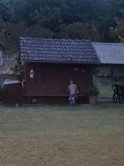 The image shows a low-angle, medium shot of a small, dark red wooden building with a tiled roof. To the left of the building is a dark gray storage container. A person, possibly a mannequin or scarecrow, is sitting on a wooden bench in front of the building. The figure is wearing a light gray long-sleeved shirt, sunglasses, and has dark hair. Blue jeans are visible around its legs. In front of the figure is a large potted plant. To the right of the building, in the background, a corrugated metal roof covers an open-air area with chairs and tables. The scene is set in a grassy area with trees in the background. There is a faint red line visible on the grass in the foreground. The lighting suggests it is dusk or early evening. The image does not provide any specific details to place it within Ludwigsburg, Germany.