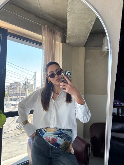 A person is captured in a mirror selfie, posing indoors with a window view of an outdoor scene. The individual, appearing to be female, is wearing dark sunglasses, a white collared long-sleeved shirt, and jeans. A colorful, patterned fabric or scarf is tied around their waist. Gold-toned bangles are visible on their right wrist. They are holding a smartphone with a leopard print case, angled to capture their reflection in the mirror. Their long, dark hair falls over one shoulder.

The indoor setting reflected in the mirror features concrete walls and ceilings, giving a somewhat industrial or modern minimalist aesthetic. A power outlet with a white cord is visible on a concrete wall section. Dark brown leather furniture, possibly chairs or a sofa, can be seen in the background of the reflection. A sheer, light-colored curtain is pulled back slightly near the window. A green plant is partially visible in the lower foreground of the image.

Through the large window, a bright, clear blue sky indicates it is daytime with sunny weather. The exterior view reveals an urban or semi-urban landscape, characterized by numerous utility poles and a dense network of overhead wires. Buildings are visible in the distance, and some sparse greenery suggests trees or bushes in the outdoor environment. The ground below appears to be paved or concrete. The overall impression is a casual, posed moment in an indoor space with an expansive view.