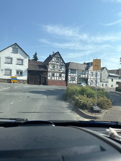 The image shows a street scene in Weidenhausen, Germany, featuring traditional half-timbered houses and a white stucco building. The perspective is from inside a vehicle, looking forward through the windshield. Two yellow directional signs are visible, one pointing left towards "Wetzlar 6 km" and "Dortmund," and another pointing right towards "Frankfurt" and "Bad Laasphe 12 km." A traffic sign displaying a U-turn symbol is also present on the right side of the road. The sky is clear blue with some wispy clouds, and the sun is shining, casting shadows on the asphalt road. Green bushes line the side of the road. The architectural style of the buildings suggests a rural or semi-rural setting within Germany.