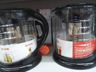 The image shows two glass kettles with black bases and handles, likely for sale on a shelf in a retail store. Both kettles have measurement markings in milliliters and cups visible on the glass. The kettle on the left has a silver metal filter inside, and the kettle on the right also appears to have a filter. Both kettles have a white band around the base with images of jars and text, likely product information or branding. A red circular seal with Chinese characters is visible on the right kettle, suggesting it has met certain quality standards. The background shows a blurry shelf or wall. The location is identified as Angren, Uzbekistan. There are no people visible in the image.