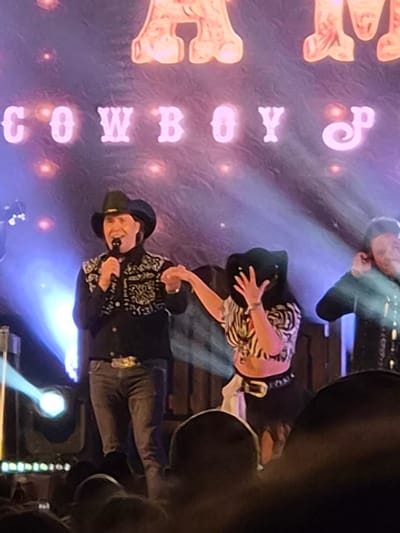 The image depicts a concert performance. Two performers wearing cowboy hats are on stage under spotlights. The backdrop includes large text reading "COWBOY" in vertically stacked letters. Stage equipment and other individuals are visible in the background. The city is not mentioned in the image.