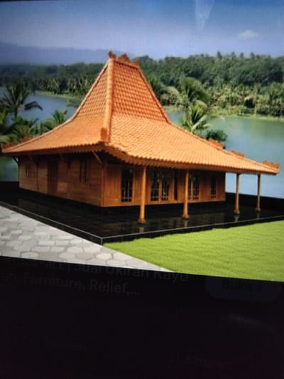 This image shows a traditional Indonesian wooden house with a tiered, orange tiled roof, typical of Javanese architecture. The house is elevated on a dark platform and surrounded by lush green grass. In the background, a body of water is visible, bordered by dense green trees and a hazy mountain range under a clear blue sky. The scene appears to be a staged photograph or rendering, likely used for promotional purposes, as indicated by text overlays at the bottom of the image which mention "artdas art | Jual Ukiran Kayu Pati, Furniture, Relief,..." and "Buka >". The specific location of Kualaraja, Indonesia is not visually depicted in the image itself, but is mentioned as a context for the scene.