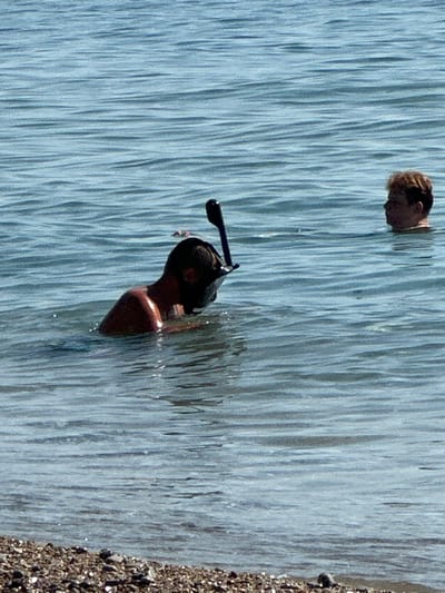 Two individuals are in the water near a shore with pebbles. The person in the foreground is wearing a snorkel mask and is partially submerged, facing to the left. Their upper body and back are visible, glistening with water. To the right and further back, another person's head and shoulders are visible above the water, facing towards the left. The environment is a body of water with visible ripples, under a clear sky. The shore in the foreground is composed of small pebbles and rocks.