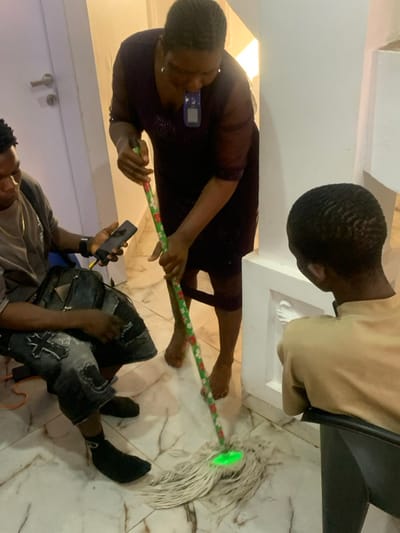 Three people are visible in the image. A woman is holding a mop with a green top. A man is sitting, holding a phone. Another man is partially visible, seated in a chair. They are inside a building with a marble floor. The mop is on the floor near the sitting man, and the woman appears to be using it. The overall lighting appears to be artificial.