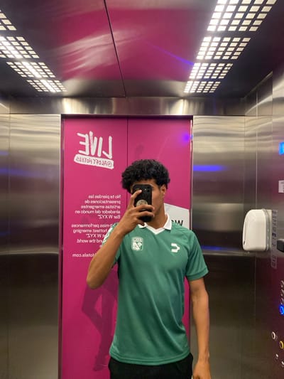 The image shows a person in a green soccer jersey taking a selfie in an elevator. The elevator has a pink accent wall with white text reading "LIVE" in a stylized font and smaller text detailing terms. The elevator features a metallic interior, illuminated by a grid of lights on the left. An elevator control panel is visible above the person, and a soap dispenser is mounted on the wall.