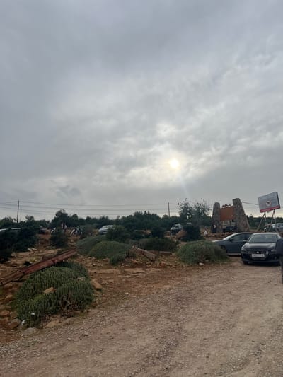 The image depicts an outdoor, sunlit scene under a partially cloudy sky, with the sun prominently breaking through the clouds in the upper left quadrant. The setting appears to be a rural or semi-rural area, possibly a hillside or mountainous region, consistent with Bin El Ouidane, Morocco.
A wide, unpaved dirt road, consisting of light brown soil and small pebbles, stretches along the right side of the frame, leading into the distance. To the left of the road, a rugged, rocky terrain is covered with lush, green vegetation, including distinctive columnar succulents and various trees and bushes.
In the mid-ground, several vehicles are parked. A dark-colored car, possibly a Peugeot, is clearly visible in the foreground with the Moroccan license plate "A4721151." Further down the road, more cars are parked, indicating a stopping point or a parking area. Near the cars and vegetation, two rustic stone pillars flank a wooden sign, forming what appears to be an entrance. Above this structure, a white sign with Arabic text displays "مرحبا بكم" (Marhaba Bikom), meaning "Welcome," along with a logo of a person swimming and other unreadable text, suggesting a tourist or recreational establishment like a cafe or camping site. Faint power lines and poles are also visible across the landscape. A few people are indistinctly seen near the parked vehicles and structures.
The time of day is clearly daytime, with the sun actively illuminating the scene through the clouds. The weather is overcast but bright, creating a diffused light. The overall atmosphere suggests an active, yet potentially tranquil, outdoor destination.