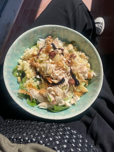 A light-blue ceramic bowl containing a salad is centrally positioned on the lap of an individual in an outdoor setting in Aldoar, Foz do Douro e Nevogilde, Portugal. The salad comprises a base of green leafy vegetables, topped with shredded cooked chicken, light-yellow couscous, and sliced cucumber. Additional ingredients include whole hazelnuts, slivered almonds, light-green pumpkin seeds, and white crumbled cheese-like pieces. A dark, viscous balsamic glaze is drizzled over the entire salad. The individual's black trousers are visible beneath the bowl. In the upper right background, a partial view of a black and white canvas sneaker is visible, featuring a white rubber sole and laces. A textured, dark-colored object, possibly a woven bag, is partially visible in the lower left foreground. The scene is illuminated by strong direct sunlight, casting distinct shadows and highlighting a reddish-brown tiled surface in the out-of-focus background, consistent with a patio or balcony.