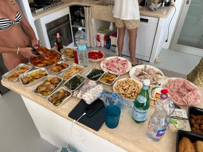 In a kitchen in Ceglie Messapica, Italy, a woman in a black and white striped bikini top and bottoms is cutting a round, tomato-topped focaccia with scissors. The focaccia is in a foil pan, and several other foil pans containing food, including what appears to be roasted chicken and potatoes, are on a countertop. There are also plates of sliced ham and other meats, a bowl of cherry tomatoes, and a bowl of bread rings. Two bottles of water, one full and one partially full, and a green bottle of Pellegrino are also on the counter. In the background, a man wearing a white t-shirt and patterned shorts is standing near a sink. A dishwasher and a stove are visible. The kitchen appears to be well-lit and modern.
