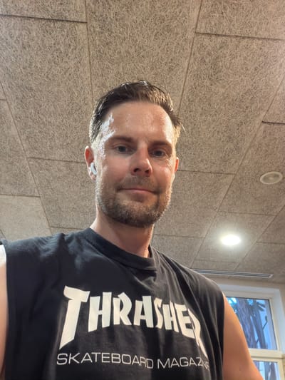 A man is taking a selfie in a room. He is wearing a black sleeveless t-shirt with the word "Thrasher" printed in white letters. He also has white wireless earbuds in his ears. The man appears to be sweating, and his hair is damp. The room has a textured ceiling with recessed lighting. The background also shows a window with trees visible outside. The image does not provide enough information to determine the location in Estepona, Spain.