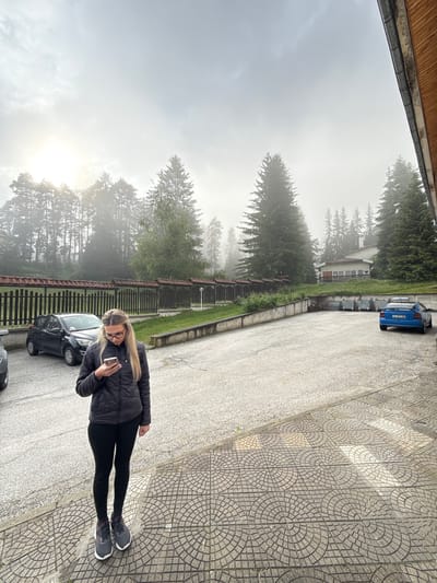 A young woman with blonde hair and glasses stands on a patterned tile walkway, looking at her phone. She is wearing a black puffer jacket, black leggings, and grey athletic shoes. Behind her is a paved parking lot with a black car parked on the left and a blue car parked on the right. In the background, a wooden fence with red roofing runs along the edge of a grassy area. Beyond the fence, a dense forest of tall pine trees rises up a hill, with a building partially visible amongst them. The sky is overcast and hazy, with the sun faintly visible through the clouds, suggesting a misty or foggy morning. The location is identified as Yundola, Bulgaria.