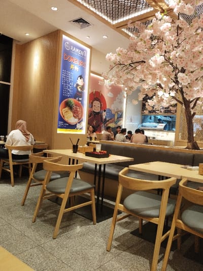 The scene is a restaurant located in Telukjambe, Indonesia, at latitude: -6.301, longitude: 107.278. The restaurant has a modern, minimalist design with wooden tables and chairs, as well as a booth seating area. A large, decorative cherry blossom tree stands in the middle of the restaurant, with branches reaching towards the ceiling.  A poster advertising "Ramen & Donburi" is prominently displayed on the wall. Several people are seated at the tables, with some looking towards the camera. Some people are interacting with each other. One woman is sitting alone at a table near the entrance, wearing a hijab. Other people are seated in the booth area, and others are standing in the background. The lighting in the restaurant is bright and warm. The floor is tiled with speckled tiles.