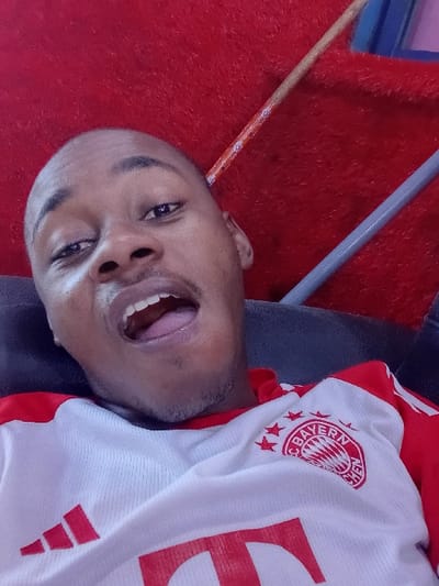 An upward-angled, close-up photograph captures an adult male in an indoor setting in Dorayi, Nigeria. The individual, appearing to be in his 20s or 30s, has dark skin, a shaved head, and light facial stubble. He is observed from the chest up, leaning against a dark, possibly upholstered surface, and looking directly towards the camera with his mouth open wide. He is attired in a white and red sports jersey, prominently displaying the FC Bayern Munich circular crest with four stars above it on the right chest, and a partial red Adidas logo on the left chest. The background is dominated by a bright red, textured fabric, likely upholstery or carpet. A light brown, rod-like object crosses diagonally from the upper left of the background, while a grey rod-like object is visible on the right side, with a small blue object partially discernible behind it.