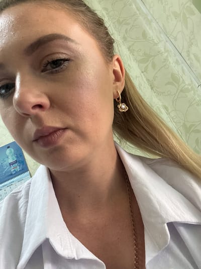 A close-up, high-angle selfie shows a young woman with blonde hair wearing a white collared shirt and a gold earring with a diamond accent. She is looking towards the camera with a neutral expression. The background is out of focus and appears to be a wall with a light-colored floral pattern. A calendar with the word "JUNE" visible is partially in the frame on the left. The lighting suggests an indoor setting. There is no information in the image to suggest it was taken in Makiivka, Ukraine.