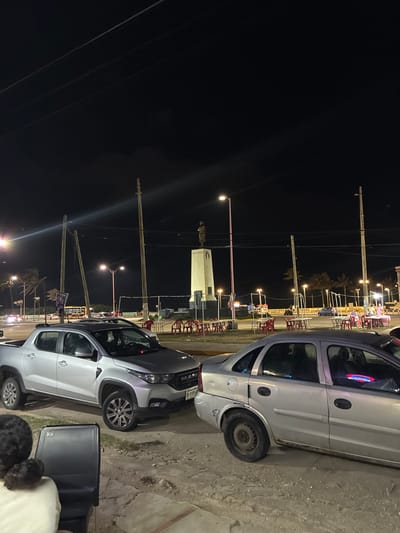 The scene depicts a nighttime urban environment in Coatzacoalcos, Veracruz, Mexico, specifically near the Monumento a Miguel Hidalgo on the Malecón Costero.
In the immediate foreground, the back of a person's head and shoulders are visible in the lower-left, seated in a black chair, facing away from the camera. Adjacent to this, a silver RAM 700 compact pickup truck is parked on a dirt and gravel surface, displaying a Veracruz license plate partially visible as "XB-8109-A." To its right, a silver Chevrolet Corsa sedan is also parked on the dirt, exhibiting visible damage on the rear passenger-side fender and a steel wheel on the front passenger side. Neon light reflections are visible in the sedan's windows.
The midground features a paved street with moving vehicles, illuminated by numerous streetlights. Utility poles with multiple overhead wires crisscross the frame. Beyond the street, multiple arrangements of red plastic chairs and small tables, some with white coverings, are distributed across grassy areas.
In the background, the prominent Monumento a Miguel Hidalgo stands tall, consisting of a light-colored, multi-tiered pedestal surmounted by a dark statue. The name "MIGUEL HIDALGO" is legible on the monument's base. Further beyond the monument, a coastal promenade or boardwalk is visible, lined with palm trees and additional streetlights, extending towards the dark horizon. The sky is dark, indicating nighttime conditions.