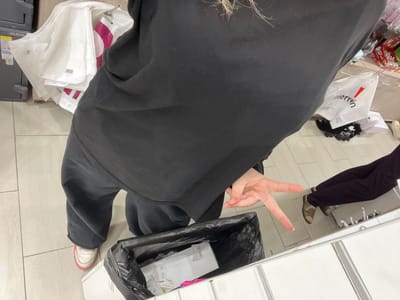 A person, viewed from a high angle, is standing in front of a metal counter and a tiled floor. The person is wearing a black sweatshirt and black pants, with white and pink sneakers on their left foot. Their right hand is raised, fingers extended in a peace sign. To the left of the person are several white plastic shopping bags. In front of the person is a black trash bin with white paper items inside. To the right, a reflection in a mirror shows the person's legs and feet, as well as another person's legs wearing dark pants and sandals. The environment appears to be an indoor retail setting, possibly a store. There is no visible indication of the location being Kokkorevo, Russia.