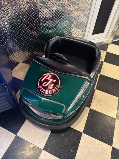 A high-angle shot depicts a green bumper car emblazoned with a "PJ's Pizzeria" logo. The logo text includes "Ocean City, MD" and "Authentic New York Style." The car sits atop a black and white checkered tile floor. The lower left side of the photo features a diamond-plated metal surface with an unknown item that is blue. The image was captured indoors. The city of West Ocean City, United States of America, is implied in the pizzeria name.