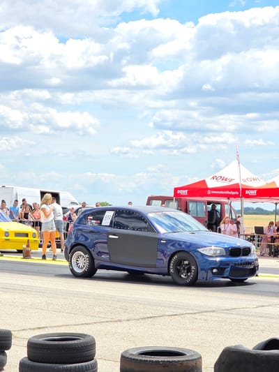 This is a vertical, medium-shot, eye-level photograph taken on a sunny day at the Akatsievo, Bulgaria drag strip. In the foreground, several stacked tires are on the asphalt. The main subject is a blue BMW with racing modifications, including a roll cage, racing tires with "Hoosier" written on them, and a black hood and door panel. The car is on a drag strip, with a yellow drag racing car to its left.

In the background, a crowd of spectators watches the event. Behind them are tents with "ROWE MOTOR OIL" branding. To the left of the tents is a red van, and beyond that, a white transit van. The sky is filled with fluffy white clouds against a clear blue backdrop. The overall scene depicts a drag racing event in Akatsievo, Bulgaria, with a focus on the modified BMW.