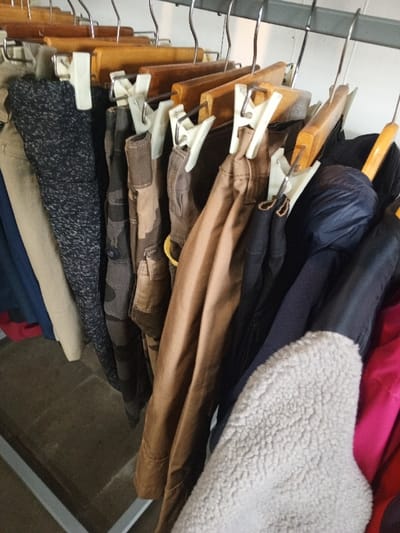 A densely packed collection of various garments hangs from a metal rod within a space in Baza, Myanmar (Burma). The items consist primarily of trousers and jackets, displayed on wooden hangers equipped with white plastic clips and suspended from a chrome-plated metal rail. Visible trousers include colors such as beige, dark blue with white speckles, camouflage patterns, dark brown, and black. Among the outerwear, a dark blue puffy jacket, a light cream-colored sherpa-textured jacket, and a magenta-colored garment are partially visible. The background is a light-colored wall or surface, and a section of a dark floor, along with a silver-colored metal frame of a clothing rack, is visible in the lower left portion of the image.