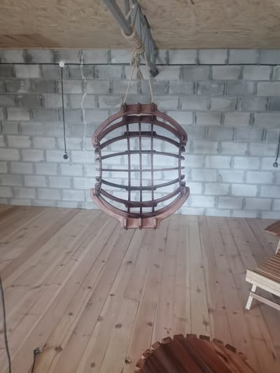 A hanging wooden sphere, composed of curved ribs and a grid-like interior, is suspended by a rope from a metal beam attached to a wooden ceiling. Below, a light-colored wooden floor extends into the scene. The background features a wall constructed of grey concrete blocks. Electrical conduits and cables are visible running horizontally along the wall. To the right, a portion of a wooden bench with slatted seating and white legs is present, and a section of a round wooden object with a serrated edge is visible in the foreground on the floor. The image appears to be taken indoors, possibly in a workshop or unfinished space, given the exposed construction materials. The scene is devoid of any people.