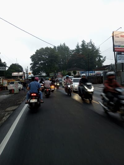 The image depicts a busy street scene in Dawathang_Dorjibi_ Kashingtsawa, Bhutan, under an overcast sky, possibly during or after rainfall. The asphalt road surface appears wet, reflecting ambient light. The scene is dominated by motorized two-wheelers, with multiple individuals traversing the thoroughfare.

In the immediate foreground, a dark-colored scooter is centrally positioned, moving away from the viewer. A person wearing a blue shirt, jeans, and a pink helmet is seated as a passenger, with their feet visible. The rider of this scooter is partially obscured.

Numerous other motorcycles and scooters follow in lanes, extending into the mid-ground. Many riders wear helmets, and some carry passengers. A white passenger car is visible amidst the motorcycles in the mid-ground, heading in the same direction. Several vehicles exhibit motion blur, particularly on the right side of the frame, indicating movement.

Along the left side of the road, a narrow unpaved shoulder or sidewalk is present. Several informal commercial structures and signs are visible, including one white stand displaying "MOTO BOX" and another sign above, partially obscured, with "HOTEL SANTO DA" text. Utility poles with an intricate network of overhead electrical or communication wires crisscross the upper portion of the frame. Lush green trees and dense foliage line the left side, extending into the background.

On the right side of the road, another large utility pole supports multiple billboards displaying various advertisements, some with Indonesian text such as "INDAH" and "BELANJA APAPUN". Beyond the billboards, more dense green trees form a natural backdrop, partially obscuring residential or commercial buildings. The sky is uniformly light grey, indicating an overcast condition and diffuse lighting.