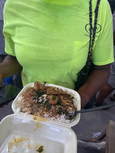 The image shows a person wearing a bright green shirt with a black chain bag strap, holding a styrofoam takeout container filled with white rice, meat stew, and dark green leafy vegetables. A black plastic spoon is in the container. The container is partially empty on one side. The person is likely in Satellite Town, Nigeria. The background is blurred and shows partial views of other people, chairs, and what appears to be a concrete surface.