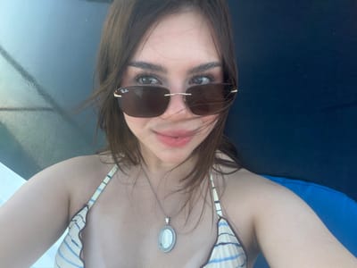 The image presents a close-up, self-portrait of a female individual in Veracruz City, Mexico, under outdoor lighting conditions. The subject, with light-brown hair, fair skin, and light-colored eyes, faces forward with a slight smile. She wears rimless sunglasses with dark lenses, featuring a "Ray-Ban" logo on the upper left lens. A silver-toned necklace with an oval pendant displaying a white cameo-style image is visible around her neck. Her attire includes a striped bikini top with light blue and beige or off-white patterns. Her hair appears to be gently moved by a breeze. The background to the left displays a bright, reflective, possibly watery or metallic surface, while the background to the right shows a dark blue, textured surface, consistent with a padded chair or outdoor furniture.