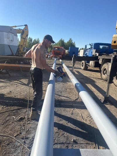 In Parma, Russia, a shirtless man wearing a black baseball cap and black pants is seen painting long pipes. The pipes are light grey with a darker grey band near the end. The man is holding a spray gun and appears to be mid-action. The scene is outdoors, with a clear blue sky suggesting a sunny day. In the background, various vehicles are visible, including a blue truck, a red truck, and a yellow excavator. The ground is rough and appears to be dirt or gravel, with shadows cast by the pipes and other objects, indicating it is likely daytime. The overall impression is one of outdoor industrial work. No visible text is present in the image.