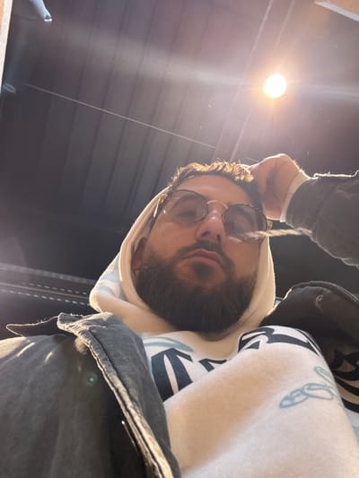 A low-angle image captures an adult male in Reichstett, France, from the chest upwards. The subject has a full dark beard and short dark hair, wearing round-framed glasses with metallic-colored rims. He is dressed in a light-colored hooded sweatshirt with light blue and dark patterns on the chest and shoulder, underneath a dark-colored, possibly denim, jacket. His right hand is visible, positioned near the back of his head. The background consists of a dark ceiling with visible linear structural elements, and a prominent bright, circular light source is visible in the upper right, causing glare on the subject's glasses and illuminating his hair. A secondary, fainter light source is also discernible in the upper left.