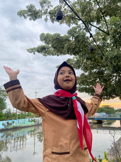 A young girl, likely of elementary school age, stands centrally in an outdoor setting in Kalitengah, Indonesia. She wears a tan-colored long-sleeved uniform shirt with brown cuffs and pocket trim, a dark brown hijab, and a red-and-white neckerchief secured with a green woggle, indicating an Indonesian scout (Pramuka) uniform. Her arms are raised with open hands, palms facing upward, and her mouth is open with a wide-eyed expression as she looks upwards and towards her left. Above her, dark-colored string light sockets are visible, strung among green leafy tree branches. In the midground, a body of water, appearing to be a canal or river, flows behind her. The far bank features a painted wall with a mural on the left and a bridge with blue and yellow railings on the right. Utility poles and vegetation are visible further in the background. The sky is overcast with grey clouds, exhibiting a warm, yellowish glow near the horizon on the right, consistent with either sunrise or sunset. The water's surface reflects the sky and elements on the bank.