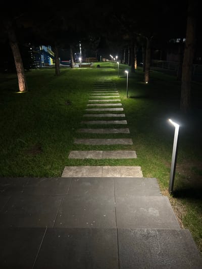 An outdoor pathway at night is illuminated by modern, minimalist pathway lights and ground lights. The pathway begins with dark, large-format tiles, then transitions to a stepping stone path laid into a manicured lawn. The lawn is flanked by tall trees, and in the background, a children's playground structure is visible, illuminated by a ground light. The scene appears to be a well-maintained park or recreational area.