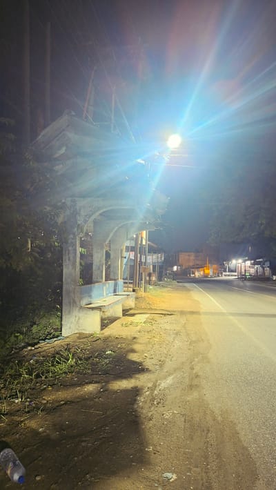 A nighttime exterior shot captures a roadside bus stop in Keudebayu, Indonesia. The bus stop features a concrete structure with arched supports and a tiled bench. A bright streetlamp illuminates the scene, creating strong lens flare and casting long shadows. The road runs parallel to the bus stop, with sparse vegetation and debris present on the roadside. Buildings and trees are visible in the background, suggesting a semi-urban or rural setting. A plastic water bottle is visible on the ground in the foreground. There are no people present in the image.