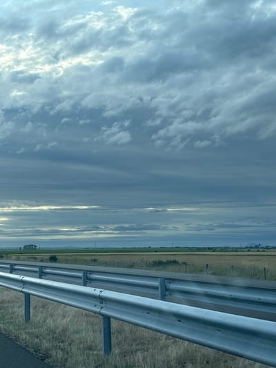 This is a photograph taken from the side of a road, looking out over a flat, rural landscape. In the foreground, a metal guardrail runs horizontally across the bottom half of the image, separating the road from a dry, grassy verge. The guardrail is silver and appears to be made of several connected sections, supported by vertical posts. Beyond the guardrail, a field of dry, golden grass or crops stretches into the distance. In the far distance, a line of green trees can be seen, and further still, a hazy horizon with faint hills. Overhead, a dramatic sky fills the upper portion of the image, characterized by a heavy layer of grey and blue clouds. Some patches of brighter sky are visible, suggesting sunlight breaking through. There are no people or animals visible in the scene. The overall impression is one of a wide-open, somewhat overcast, rural environment, likely taken from a moving vehicle due to the perspective of the guardrail. The location of Bozduganovo, Bulgaria is not indicated in the image itself.