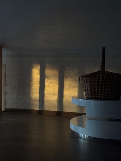 The image depicts an interior scene, likely a room or hallway, in the city of Dubki, Russia. The perspective is low-angle, looking towards a light-colored wooden plank wall at the rear. Direct sunlight enters from an unseen source to the left, casting three vertical rectangular patterns of yellow-orange light and associated dark shadows onto the wooden wall. The floor is dark and uniform, possibly a dark wood or laminate. In the right foreground, a dark brown, woven basket with a prominent handle rests on a multi-tiered, white, circular table. The table has at least two visible tiers, with the upper tier supporting the basket and appearing smaller in diameter than the lower tier. A subtle metallic trim is visible at the base of the table. The overall environment is dimly lit outside the sunlit areas, creating a high-contrast scene. No individuals are present.