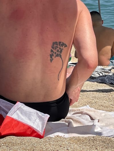 The image shows the back of a man with a tattoo of a manta ray on his right side. He is wearing black swim trunks. In front of him, partially visible, is a red and white mesh bag. A white towel is laid out on the sand behind him, and a portion of another person's back and head, also facing away from the camera, is visible in the background, seated on the sand. The scene appears to be a beach, with sand in the foreground and the ocean visible in the background. The water is blue and calm. The lighting suggests it is daytime. There is a slight reddish hue on the man's back, indicative of sunburn. This image was taken at a beach in Badalona, Spain, a coastal city in Catalonia known for its beaches.