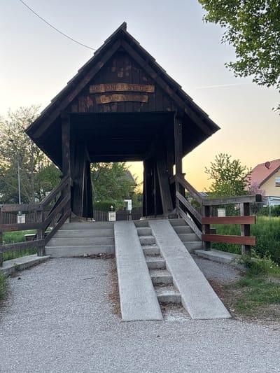 The image shows a wooden bridge structure, likely a pedestrian overpass or bridge, known as the "19. Pionier-Steg" in Wiener Neustadt, Austria. The bridge has a dark-stained wooden frame and roof, with wooden railings and supporting beams. There are concrete stairs leading up to the bridge on both sides, with concrete ramps positioned alongside the stairs. The environment includes green trees and vegetation on both sides of the bridge. A building can be seen in the background. The image is taken during sunset/dusk, with a warm glow. The path leading up to the bridge is asphalt.