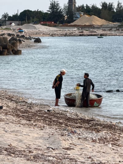 Two men stand in shallow water next to a round, red basket-like boat on the shore of Lý Sơn Island, Vietnam. The man on the left wears a black t-shirt with yellow writing and black shorts, with a white cap on his head. He faces the other man with his hands behind his back. The man on the right wears a black long-sleeved shirt and black pants. He holds a fishing net with yellow items in it, and it is draped over the boat. The boat is partially submerged in the water. The shore is a mix of sand, pebbles, and dried seaweed. In the background, a rocky breakwater made of concrete tetrapods is visible on the left, and the sea extends to the horizon. Further inland, there is a sandy hill with construction materials and buildings. The sky is overcast.