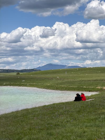 In the city of Javorje, Montenegro, a panoramic outdoor scene features a serene body of water, expansive green hills, and a dramatic sky. The upper portion of the image is dominated by a blue sky with large, white cumulus clouds, interspersed with some darker grey cloud formations. The middle ground consists of undulating green hills, densely covered with grass, with scattered white rocks and a small, isolated tree visible on the left side. Distant mountains rise in the background, with one peak showing a lighter patch near its summit.

In the lower portion of the image, a calm, light green or turquoise body of water extends from the lower left towards the center. On the grassy bank bordering this water, in the lower right, two individuals are seated with their backs to the viewer. The person on the left is wearing a black jacket, while the person on the right is wearing a bright red jacket. They are positioned close together, facing the water. A colorful blanket is visible on the grass to the right of the person in red. The immediate foreground also shows green grass interspersed with small blue and yellow wildflowers. A vertical, rectangular marker is visible on the hillside in the mid-ground, slightly to the left of the seated individuals.