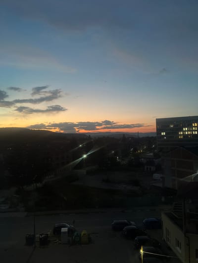 The image captures a twilight view of the city of Samokov, Bulgaria, with the sky displaying a gradient of blue to orange hues from sunset. In the foreground, a street is visible with several parked cars and a row of trash bins. Buildings, including a large multi-story structure with illuminated windows on the right, are present in the mid-ground and background, interspersed with trees and darker, silhouetted structures suggesting construction or undeveloped areas. The horizon is marked by a line of hills or mountains, partially obscured by trees and the darkening landscape. Streetlights illuminate parts of the scene, adding to the ambiance of early evening.