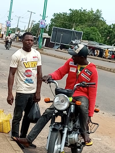 The image depicts a street scene in Garko, Nigeria, where two men are standing near a motorcycle. The man on the left, wearing a light-colored t-shirt with "VIBES" printed on it in yellow, and dark pants, stands facing forward with his hands at his sides. He is holding a dark plastic bag in his left hand. The man on the right, seated on the motorcycle, is wearing a red hooded jacket with multiple patches and a cap. He is looking to his left, with his right hand resting on the motorcycle's handlebars. The motorcycle is black with chrome accents and an orange turn signal visible. In the background, a yellow bag is on the ground to the left, and a blurry image of another motorcycle with a rider is seen in the distance on the road. Other elements in the background include street poles with posters, trees, and buildings, suggesting an urban environment. The overall scene appears to be a casual street interaction or stop.