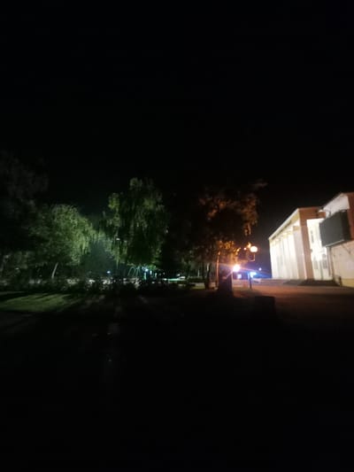 The image depicts a nighttime scene in Parvomay, Bulgaria, focusing on a building with classical columns, likely a public institution or cultural center. The building's facade is illuminated by artificial lights, highlighting its architectural features. In the foreground, to the left, are several trees with lush foliage, also partially lit, casting shadows on the ground. A street lamp with an orange glow is visible between the trees and the building. The overall impression is that of a well-maintained public space at night, with the building serving as a prominent landmark. The darkness of the sky and the selective illumination suggest the photograph was taken after sunset, possibly in the evening or late at night. There are no people visible in the frame, and the environment appears serene and empty.